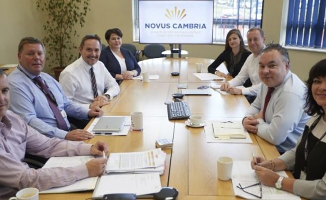 Novus Cambria team in a meeting room, discussing learning and skills development, with a Novus Cambria logo displayed on a screen.
