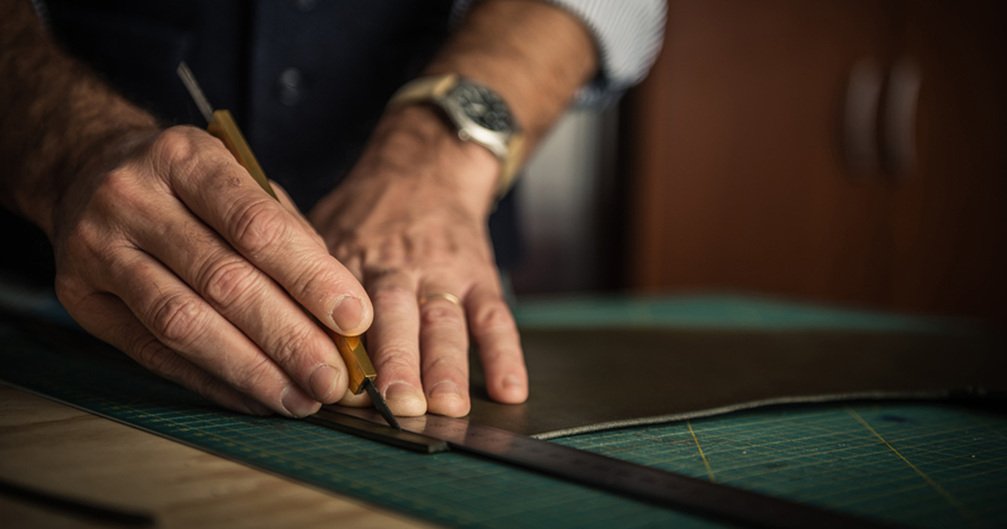 Leather worker using a craft knife and metal ruler to cut brown leather on a gridded surface.