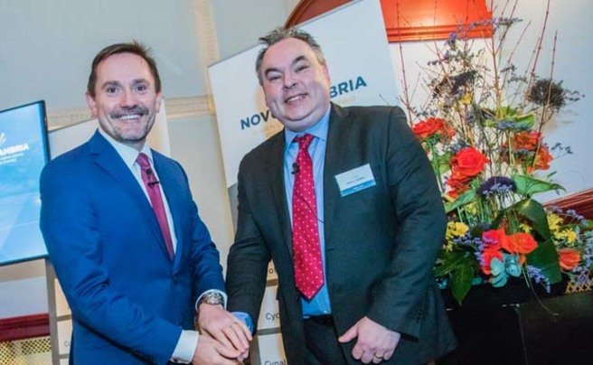 Two men in business attire shaking hands at a Novus Cambria event, celebrating a partnership, with a branded backdrop.